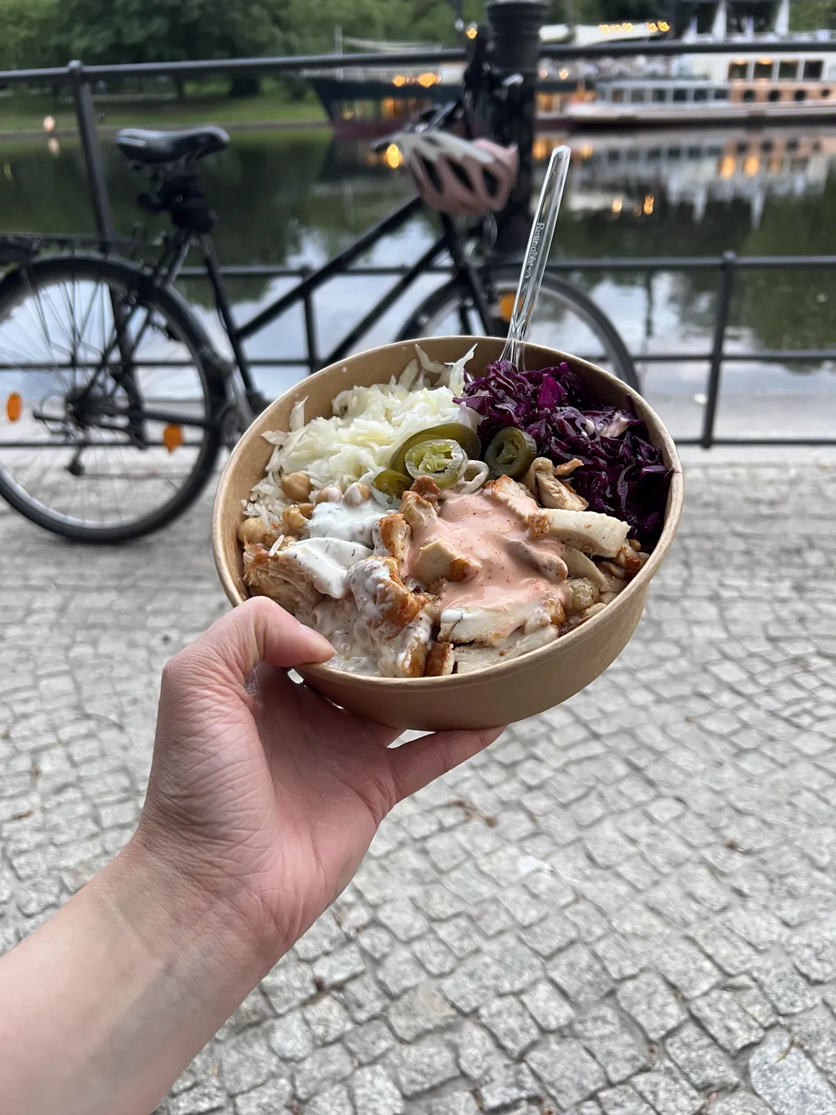 Eine Hand hält eine Chicken Bowl am Landwehrkanal, im Hintergrund Fahrrad und Wasser