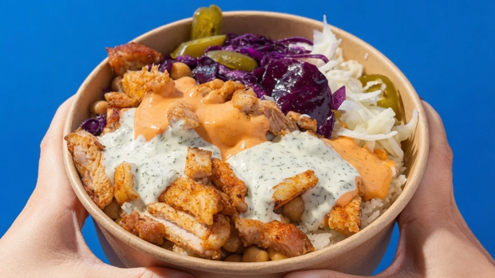 Nahaufnahme der Chicken Rice Bowl im Kraft-Karton: Reis, gebratenes Hähnchen, Kichererbsen, Rotkohl, Krautsalat, Jalapeños und zwei Saucen — links und rechts halten Hände die Bowl
