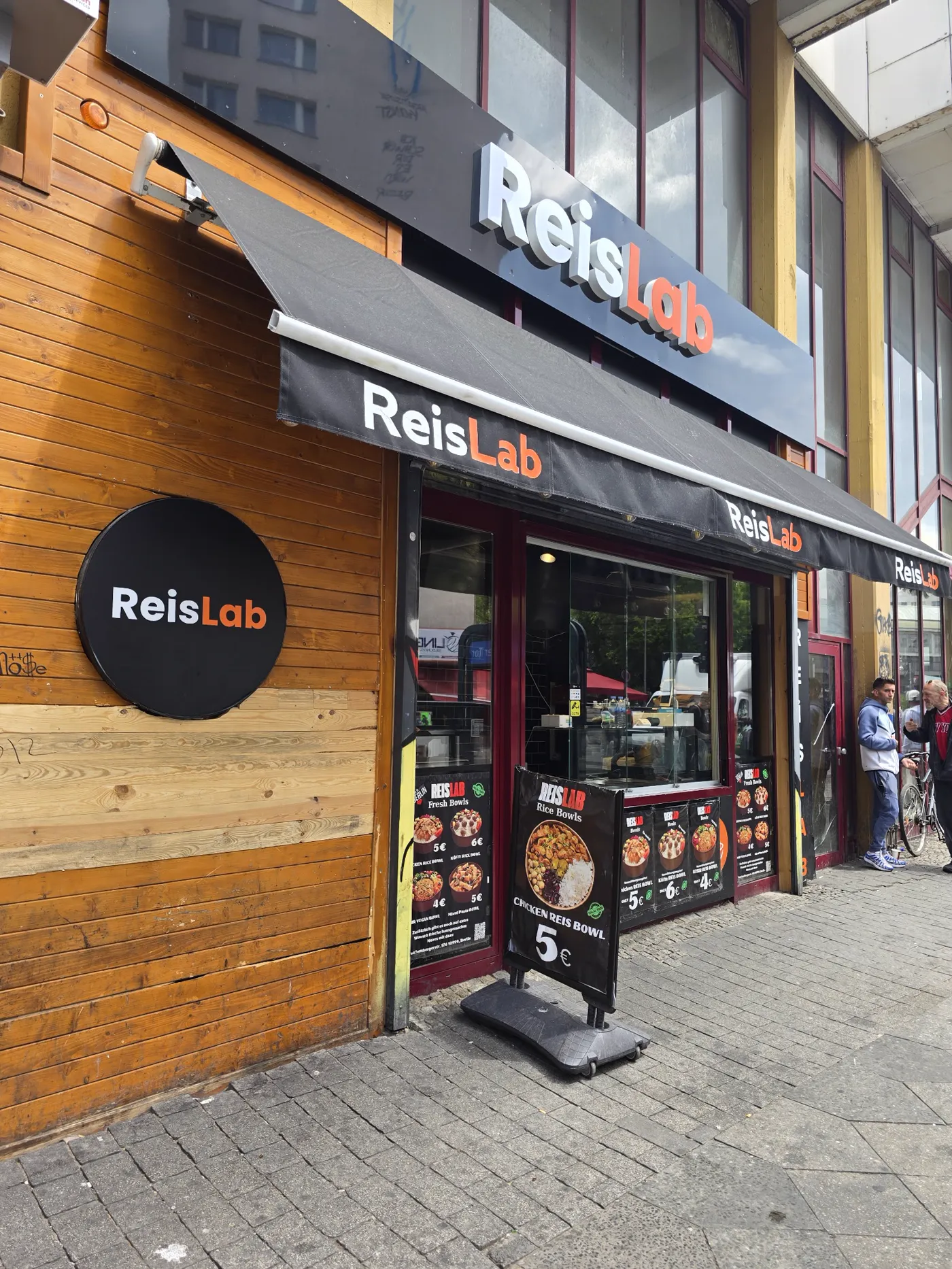 Ladenfront von ReisLab Bowls: Holzfassade, schwarze Markise mit orangefarbenem Logo und Menü-Plakate im Fenster